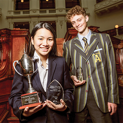 A young woman smiling and holding a trophy and a young man pictured in the reading room of the state library  victoria