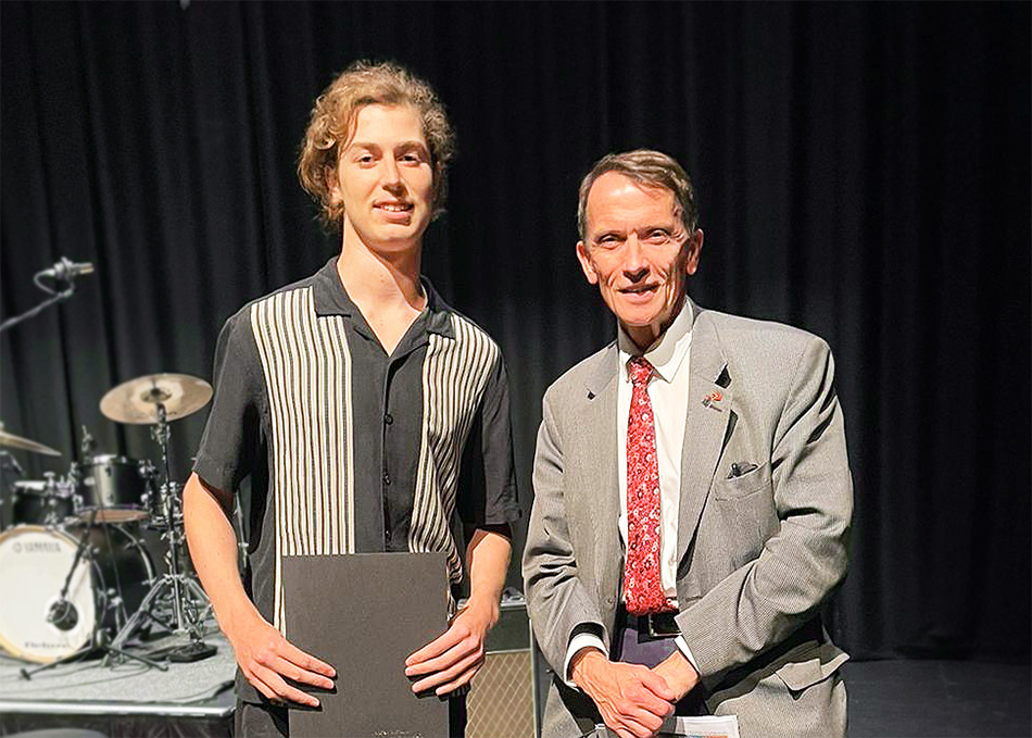 Two people stand on a stage shoulder to shoulder and smiling. The person on the left is holding a black folder.