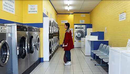 A young woman in a red coat stands in a colourful Laundromat with a blank expression