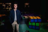 A person with brown hair wears a light blue shirt, khakis, and a navy-blue jacket. He stands next to a meter tall wooden Rubik’s cube that is mid rotation.
